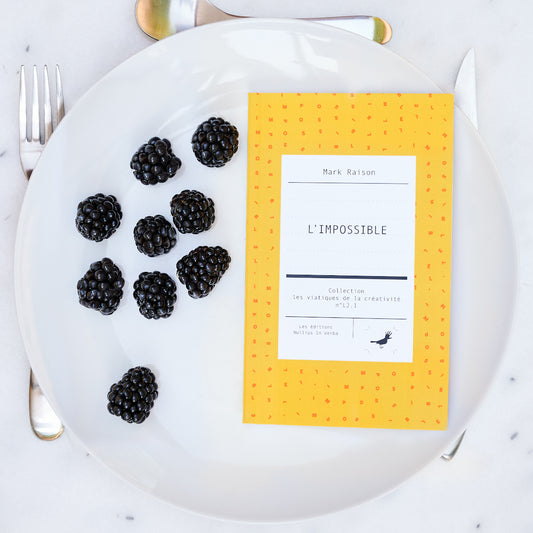 Le livre est disposé dans une assiette avec des mures. On voit une fourchette, un couteau et une cuiller autour de l'assiette.