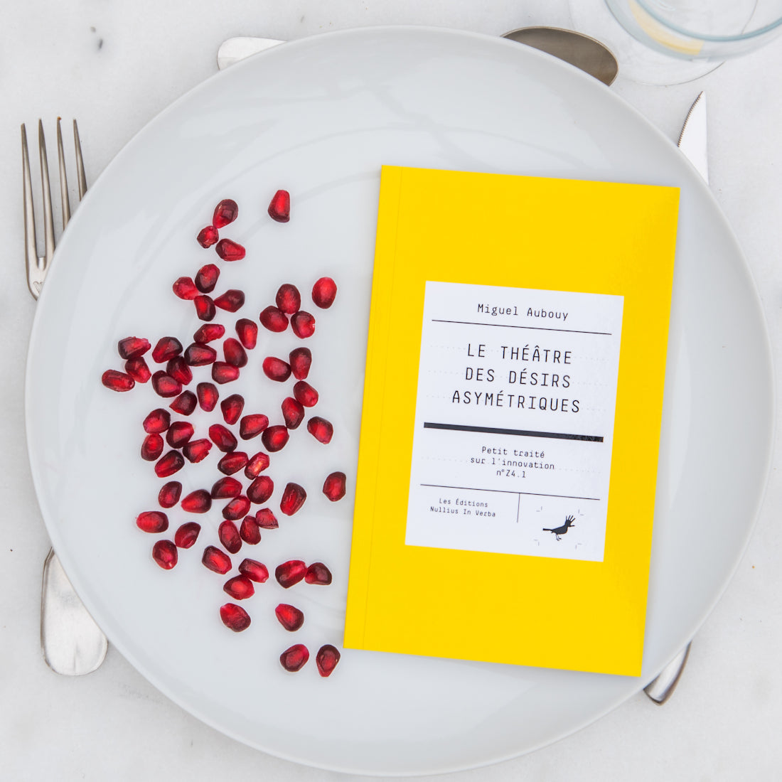 Le livre est disposé dans une assiette avec des grains de grenade. On voit une fourchette, un couteau et une cuiller autour de l'assiette.