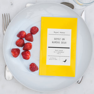Le livre est disposé dans une assiette avec des fraises. On voit une fourchette, un couteau et une cuiller autour de l'assiette.
